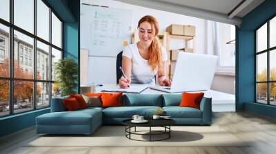 Young female business owner sitting in office writing sales and deliveries on paper sheets while using laptop Wall mural