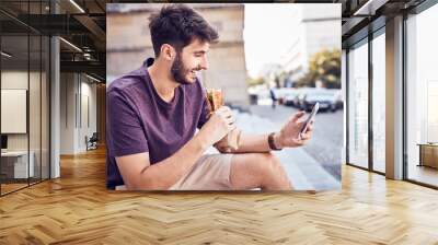 Young man eating lunch and using phone outdoors in the city Wall mural