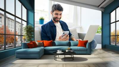 Young man working at home using laptop and smartphone Wall mural