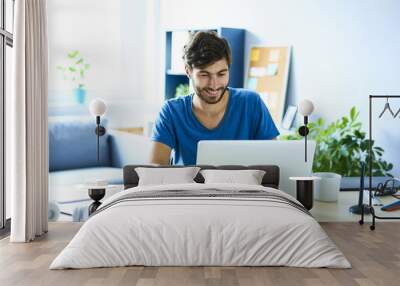 Young man working on laptop in home office and smiling Wall mural