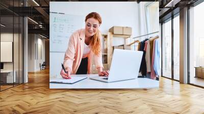 Young woman marking deliveries on clipboard and looking at laptop in office Wall mural