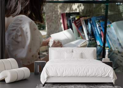 a young girl reads an open book near a glass rack, many books in multi-colored binding on bookshelves, rear view, natural lighting Wall mural