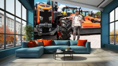 Caucasian adult man with digital tablet in hand stands next to construction or agricultural tractor at a machinery dealer. Ag equipment dealer or supplier. Wall mural