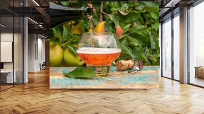 Pouring of rose apple cider from Normandy in glass, France and green apple tree with ripe red fruits on background Wall mural