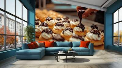 Close shot of many creamy profiteroles on the foreground while a baker drizzles chocolate on the last one Wall mural