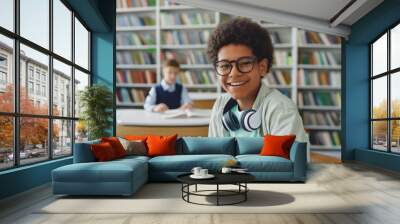 Happy young black teenager is in the library, studying for exams, while wearing headphones around his neck. Wall mural