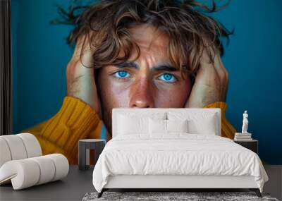 Distressed Young Man with Disheveled Hair Clutching Head in Agony, Expressing Intense Headache or Migraine Pain on Blue Background Wall mural