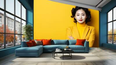 Thoughtful Asian businesswoman in a yellow turtleneck and blazer standing with crossed arms against a matching yellow background, exuding confidence and professionalism Wall mural