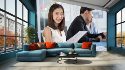 Attractive asian young confident businesswoman sitting at the office table with group of colleagues in the background. Business woman with coworkers in meeting room. Wall mural