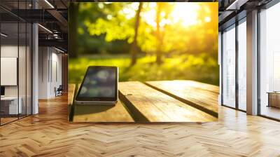 A mobile phone rests on a wooden table in a park, with the sun shining brightly in the background. Wall mural