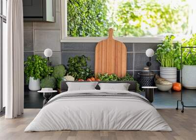 Fresh vegetables and herbs arranged on a kitchen counter with a wooden cutting board and potted plants in front of a window. Wall mural