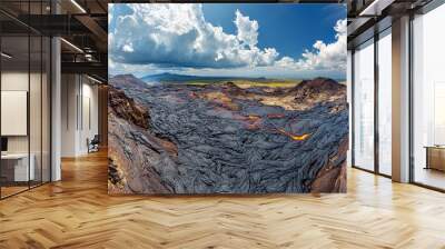 Panoramic view of a volcanic landscape with solidified lava flows, glowing red lava streams, and a blue sky with puffy clouds. Wall mural