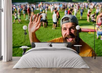 Hipster in cap visiting social event picnic fest or festival. Urban event celebration. Man waving hand sunny day outdoors. Man bearded hipster in front of crowd people green field background Wall mural