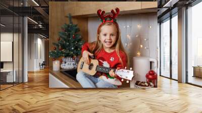 Happy little girl in red horns playing ukulele on the background of christmas tree. Talent kid having fun on winter holidays at cozy decorated home kitchen Wall mural