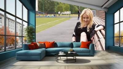 The girl sits on the bench at the football field with the ball. Wall mural