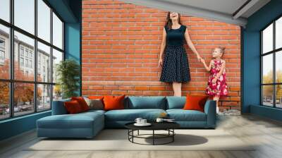 Fashion family concept - stylish mother and child. A portrait of a happy family: a young beautiful woman with her little cute cheerful daughter standing near brick wall. Wall mural