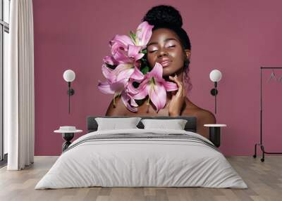 Fashion beauty portrait of young African American model with pink art make up posing with lily flowers against pink background. Wall mural