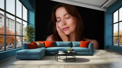 Softness. Skincare model with brown hair is touching her face and posing against black background Wall mural