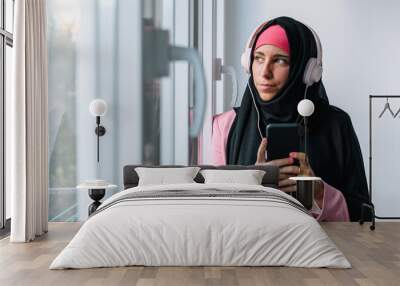 Young muslim woman wearing a hijab using technology indoors. Horizontal side view of arabic woman listening to music with smartphone next to a window at home. Technology and muslim women lifestyle. Wall mural