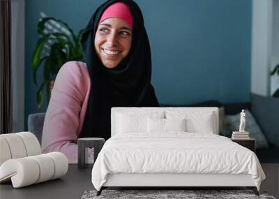 Young muslim woman wearing a hijab using technology indoors. Horizontal side view of arabic woman working with smartphone at home. Technology and muslim women lifestyle. Wall mural