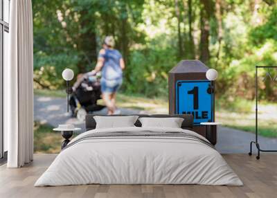 A one mile marker sign post beside a paved pathway with a bright green and sunny background with a mom walking and pushing a stroller. Wall mural