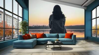 Back view of long hair brunette girl sitting on the pier near lake at sunset time. Wall mural