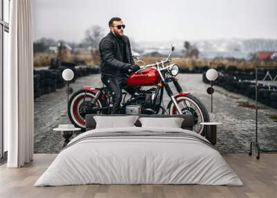 Red motorbike with rider. A man in a black leather jacket and pants stands sideways in the middle of the road. Tires are laid on the background Wall mural