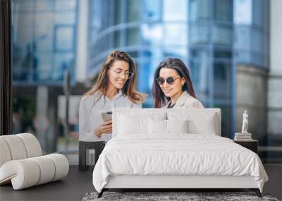 Two young business lady with tablet and notebook walking outdoors near modern business center. Wall mural