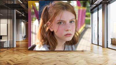 A beautiful teenage girl with ponytails and freckles flushed from the heat. Red-haired girl with a pensive, serious look looks straight, sitting on a bench in an amusement park in summer. Wall mural