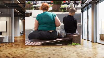 A fat overweight mother and her slender son sit and relax on a bench in a summer European city. Back view of passers-by, lifestyle. 07/15/2020. Wall mural