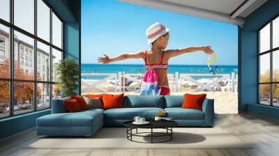 Back view of girl stands on sandy beach with sand toys and swimming goggles and looks at blue sea. Vacation summer travel holiday Wall mural