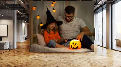 Father and little daughter in witch black hat watch horror film on tablet in dark with orange pumpkin lamp. Halloween celebration Wall mural