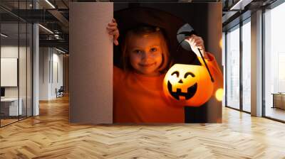 Halloween. Smiling little girl in witch costume and black hat look out from corner with lantern pumpkin. Wall mural