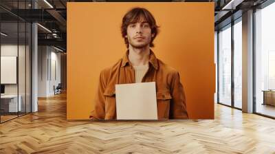 Young man holding blank poster against vibrant orange backdrop, serious expression, casual attire Wall mural