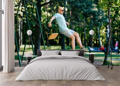 Odd adult bald man in sunglasses jumping from swing on ground in motion.  Childhood behavior. Hulking awkward foolish comic male have fun on children playground outdoor in city park in summer day. Wall mural