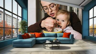 Young happy mother feeding her little baby with delicious tasty soup from plate in kitechen. Funny portrait of eating child. Beautiful girl in glasses together with daughter have diinner at home. Wall mural