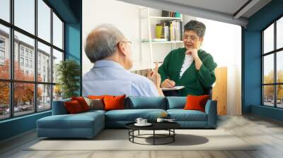 Depressed Asian senior man sharing problems during therapy session with female psychiatrist writing on clipboard. Wall mural