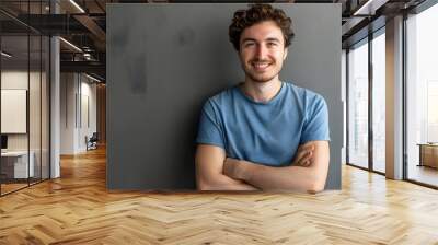 Indoor portrait of young european caucasian man isolated on gray background, standing in blue t-shirt with crossed arms, smiling and looking straight at camera.  Wall mural