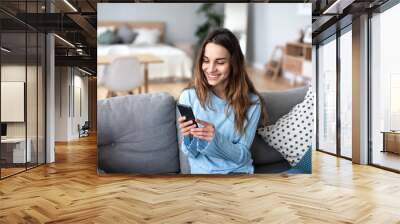 cheerful young woman using mobile phone while sitting on a couch at home. Wall mural