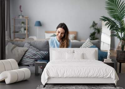 Online chat. Smiling woman talking on smartphone with friends while sitting on sofa at home. Wall mural