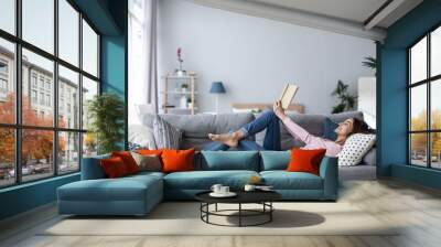 Young beautiful woman is reading a book while lying on a sofa in the living room. Wall mural