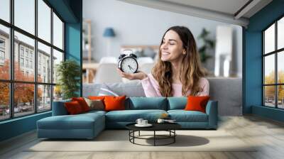 Young smiling woman holds in hand alarm clock on the couch In a living room Wall mural