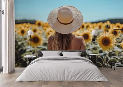 A woman with a wide-brimmed straw hat standing in a sunflower field, facing away, embracing the summer vibes, with clear skies above. Wall mural