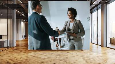 Businesswoman holding digital tablet and looking at handsome colleague while shaking hands in office Wall mural