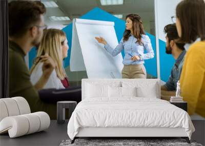 Confident young team leader giving a presentation to a group of young colleagues as they sit grouped by the flip chart in the office Wall mural