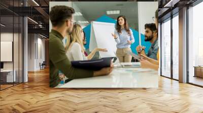 Confident young team leader giving a presentation to a group of young colleagues as they sit grouped by the flip chart in the office Wall mural