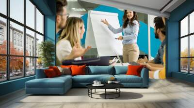 Confident young team leader giving a presentation to a group of young colleagues as they sit grouped by the flip chart in the office Wall mural