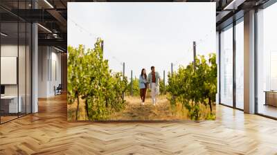 Couple strolls hand in hand along a sunlit path amidst lush vineyards during a warm afternoon Wall mural