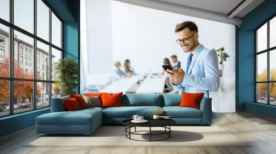 Happy young man using his mobile phone and smiling while his colleagues working in the background Wall mural