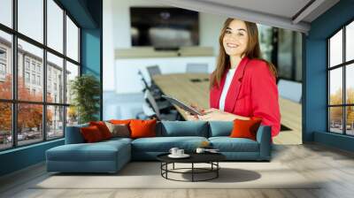 Smiling professional woman in red blazer holding tablet in modern conference room during daytime Wall mural